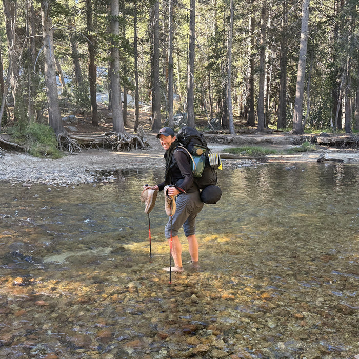 A hiker stands in a shallow stream holding a fish, with a large backpack and hiking poles, his feet clad in rugged MACV-2 - Mid Top - Briar + Coyote boots, surrounded by trees.