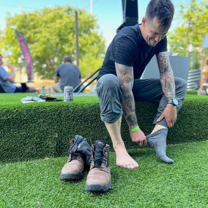 A man sits on grass, putting on socks beside his MACV-2 - Mid Top - Chestnut + Black boots with a can nearby and others in the background.