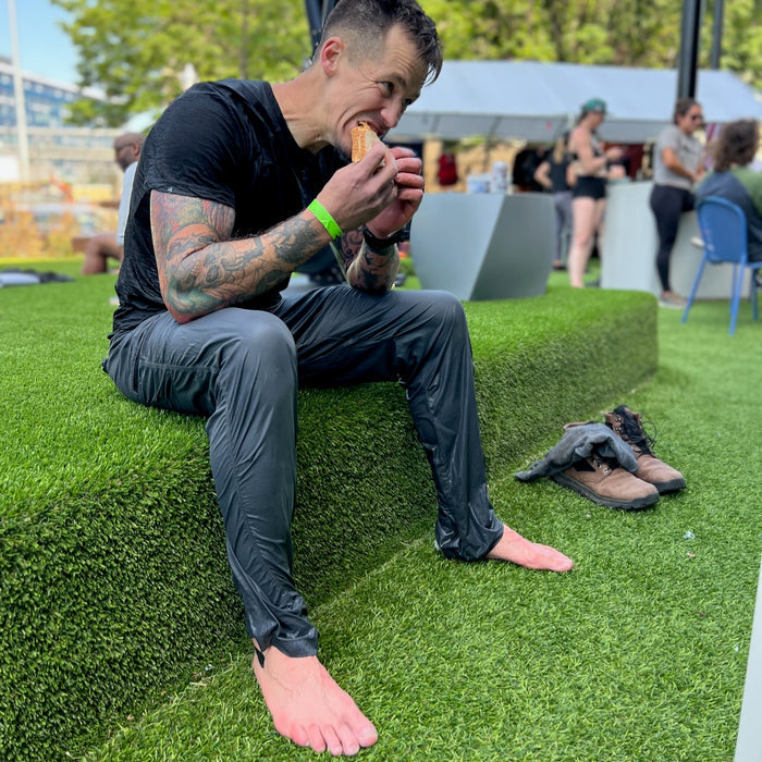 A man in wet clothes sits barefoot on artificial grass, eating a sandwich with his MACV-2 - Mid Top - Chestnut + Black boots and gloves beside him, the aggressive outsole of his Special Forces boots visible.