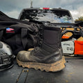 The MACV-2 Safety Boot - 8" High Top sits muddy and black on a truck bed, surrounded by work gear, a helmet, and a Stihl chainsaw in the background.