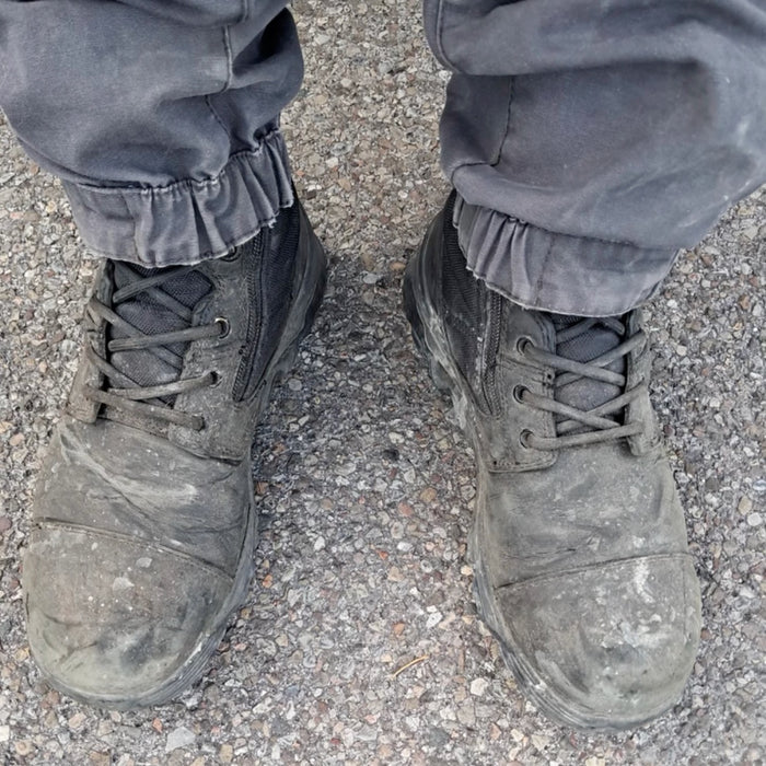 Wearing the MACV-2 Safety Boot - 8" High Top in black, paired with gray pants, standing on a gravel surface as seen from above.
