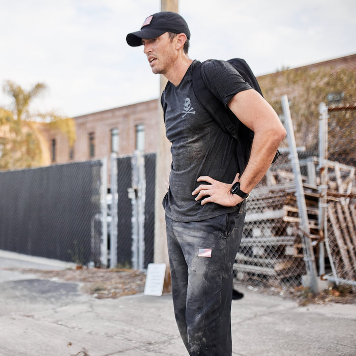 A man stands outside near a fence, wearing a cap, a dusty black shirt, and tough Men’s American Training Sweatpants made in the USA, with a backpack on his shoulder.