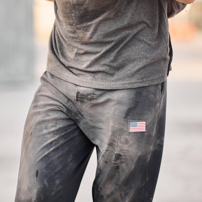 Wearing the Men’s American Training Sweatpants—durable, dirt-stained gray with a small American flag patch—and a matching dirty gray t-shirt, proudly made in the USA.