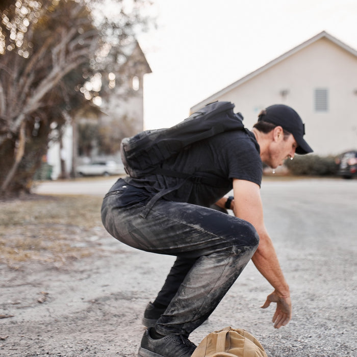 A person wearing dark clothes and Men’s American Training Sweatpants with a backpack crouches on a gravel road, reaching toward a tan bag on the ground.
