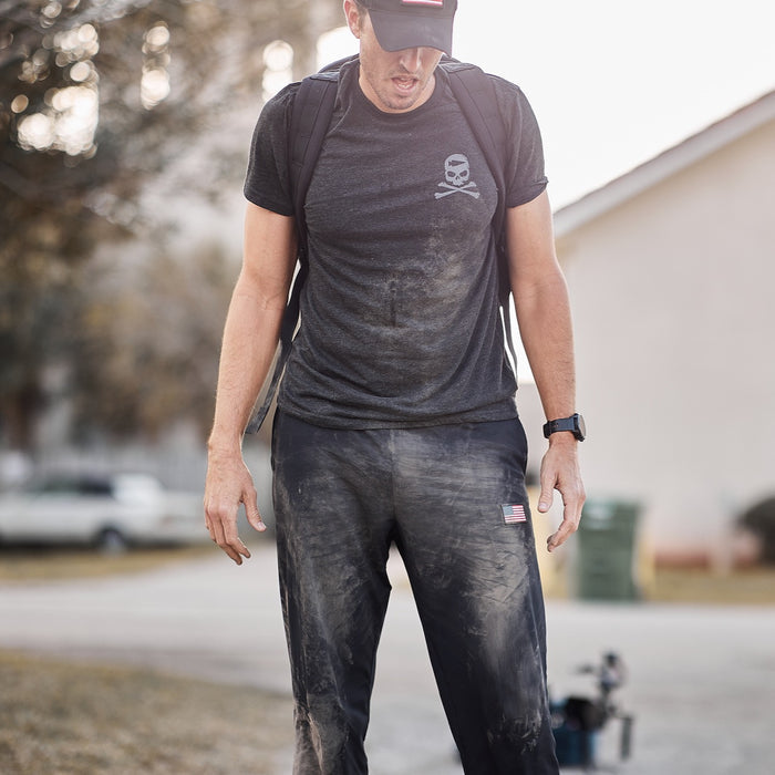 A man in dirty clothes and a backpack stands outside, looking down, with dirt on his shirt and pants—his Men’s American Training Sweatpants, made in the USA and backed by a lifetime guarantee, bearing the marks of a long day outdoors.