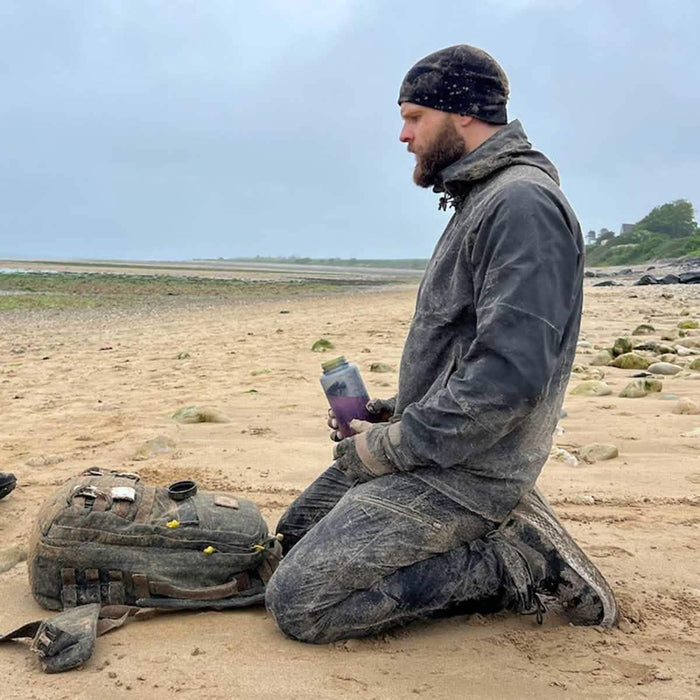 Man kneeling on sandy, rocky beach with muddy GORUCK backpack and water bottle, outdoor gear