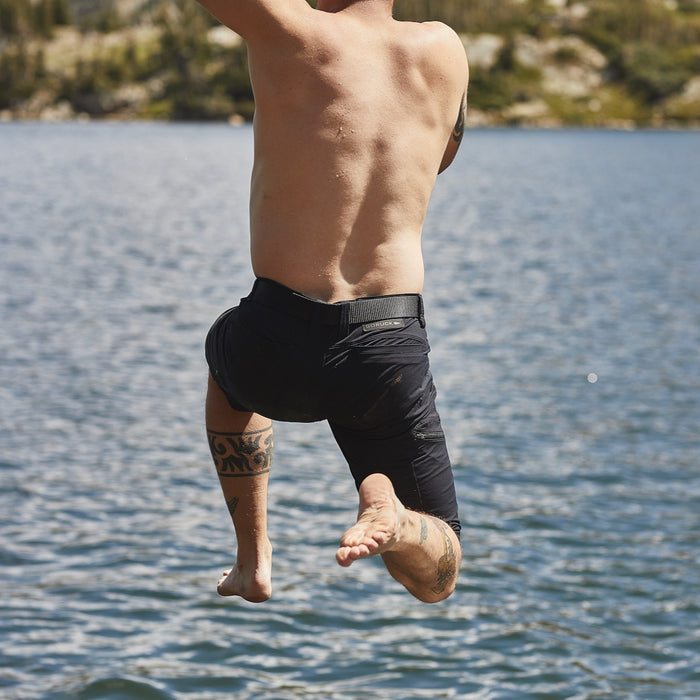 A shirtless, tattooed person jumps into a lake with water and trees in the background, wearing Men’s Challenge Shorts - Lightweight ToughDry®, abrasion-resistant technical shorts built for rugged performance.