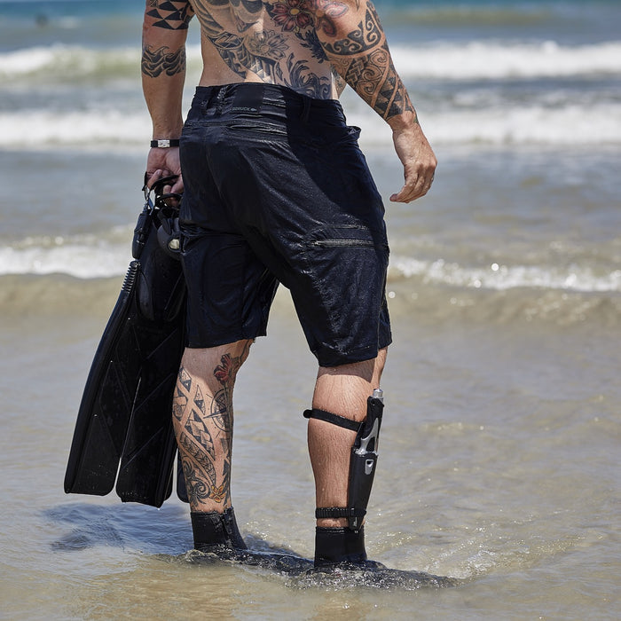 A tattooed man with a prosthetic leg, wearing Men’s Challenge Shorts - Lightweight ToughDry®, holds swim fins while standing in shallow ocean water facing the waves.