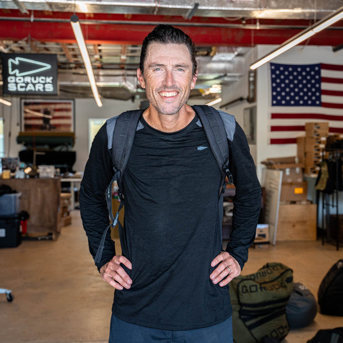 A man stands with his hands on his hips, wearing the Men’s Commando Long Sleeve - Merino Wool shirt and a backpack, in a workspace decorated with American flags.