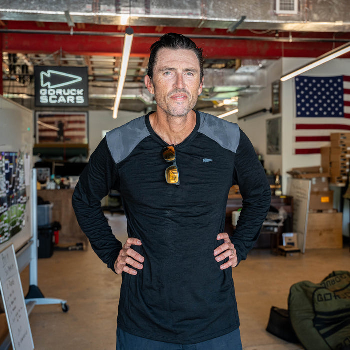 A man wearing the Men’s Commando Long Sleeve - Merino Wool shirt stands in a workshop with hands on hips; an American flag and GORUCK sign are visible in the background.