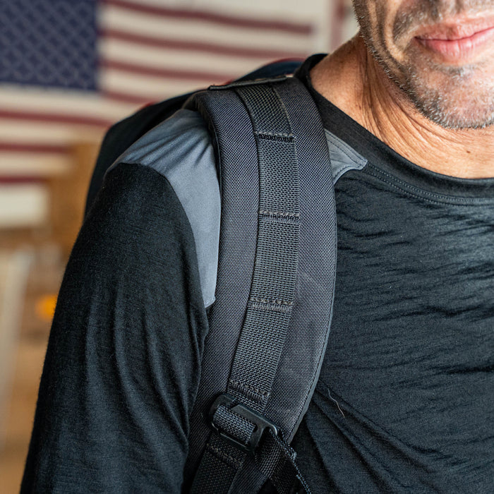 A man wearing the Men’s Commando Long Sleeve - Merino Wool shirt and a backpack stands in front of a blurred American flag.