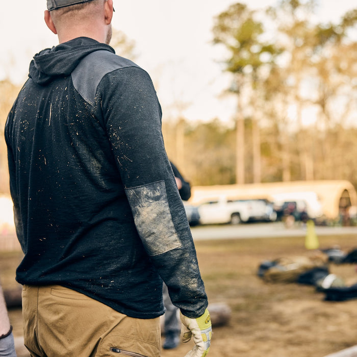 A person in dirty work clothes and gloves stands outdoors near parked vehicles and scattered equipment, wearing the Men’s Commando Pullover - Merino Wool.