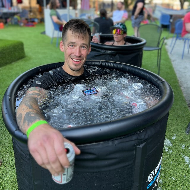 Smiling man outdoors in an ice bath, holding a drink and wearing the Men’s Commando Tee - Merino Wool; another person in a similar tub is behind him.