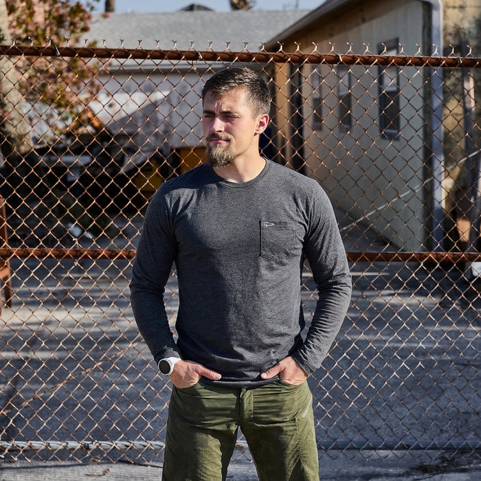A man wearing the Men's Long Sleeve Pocket Tee - Tri-BlendX in gray stands in front of a chain-link fence on a sunny day.