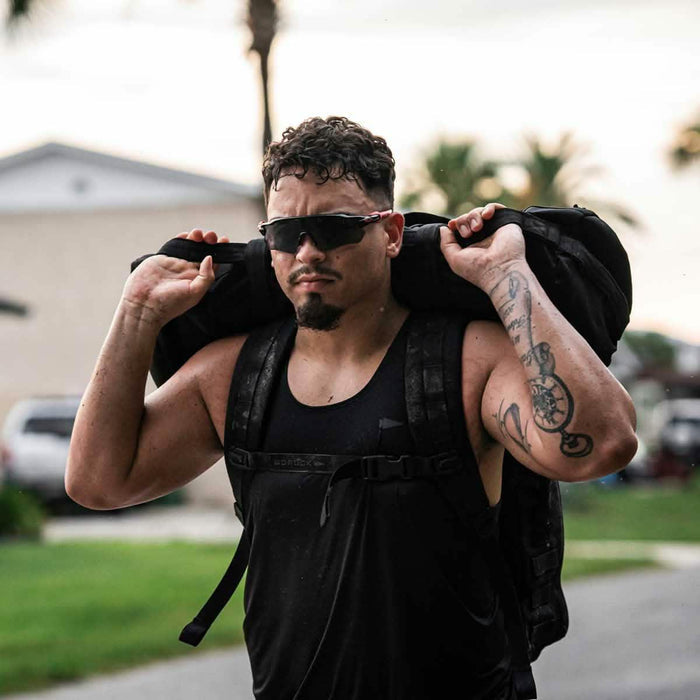 Man in black GORUCK performance tank carrying rucksack outdoors, wearing sunglasses