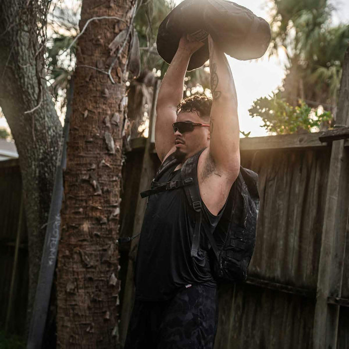 Man in GORUCK black performance tank lifting sandbag outdoors, wearing backpack and sunglasses