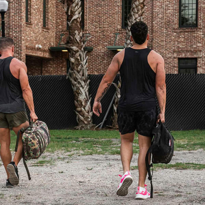 Two men in GORUCK black performance tanks walking outdoors with rucksacks, brick building background