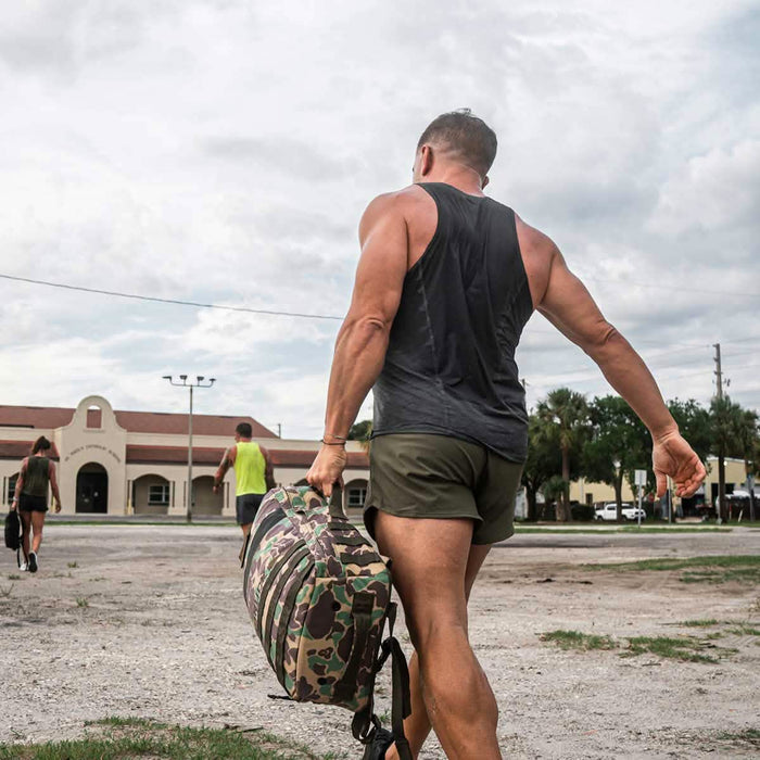 A man in athletic wear and a Men's Performance Tank - ToughMesh carries a camouflage duffel bag outdoors, walking toward a building with others.