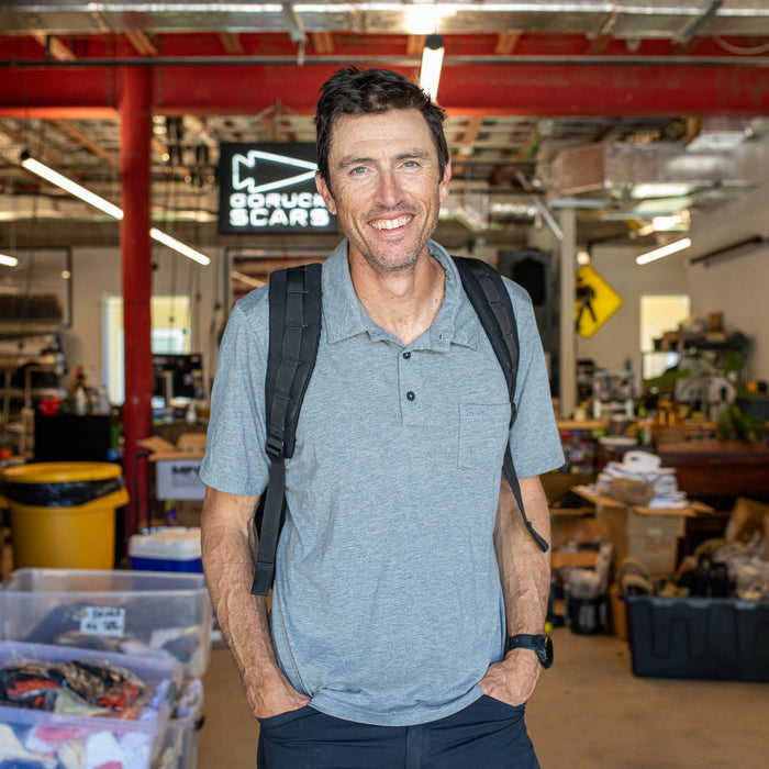 A man wearing the Men's Polo with Pocket - Tri-BlendX, featuring anti-stink fabric, stands smiling with a backpack in a cluttered workshop or garage.
