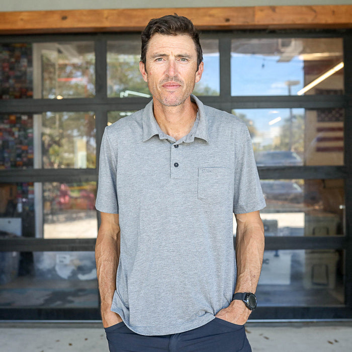 A man stands outside in front of glass doors, wearing the Men's Polo with Pocket - Tri-BlendX, hands in pockets, highlighting the style and comfort of its premium tri-blend fabric.