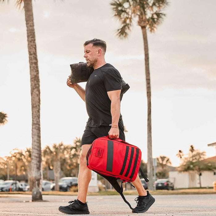 Man rucking outdoors with a red GORUCK backpack and training sandbag, palm trees in background
