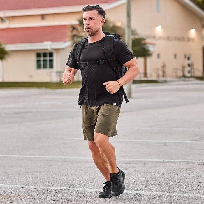 Man rucking outdoors wearing GORUCK backpack, black shirt, and olive shorts in a parking lot.