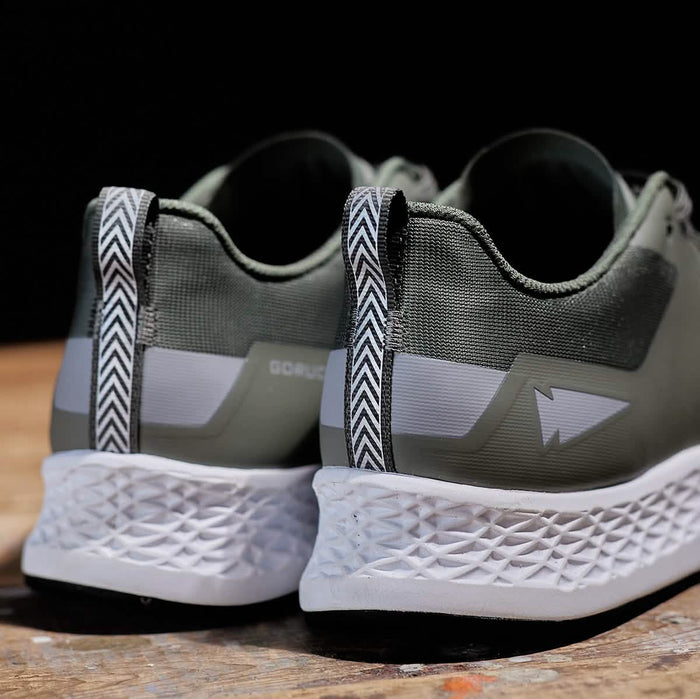 The Men's Rough Runner - Earth by GORUCK highlights intricate green athletic detailing and features textured white soles. A chevron-patterned pull tab enhances the heel, while a bold white lightning bolt embellishes the side. Set against a rustic wooden background, these shoes perfectly embody the Rough Runner spirit.