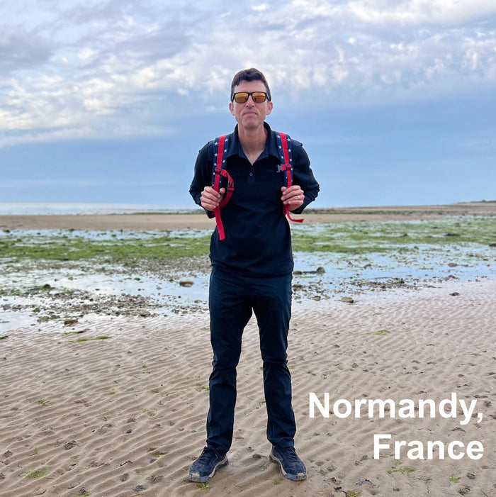 Man wearing Men's Rucking Shirt - Merino Wool in black with a red backpack stands on a sandy beach under cloudy skies. Text: "Normandy, France.