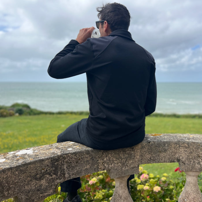 Wearing the Men's Rucking Shirt - Merino Wool, a man sits on a stone fence, drinks, and gazes at the sea beneath a cloudy sky.