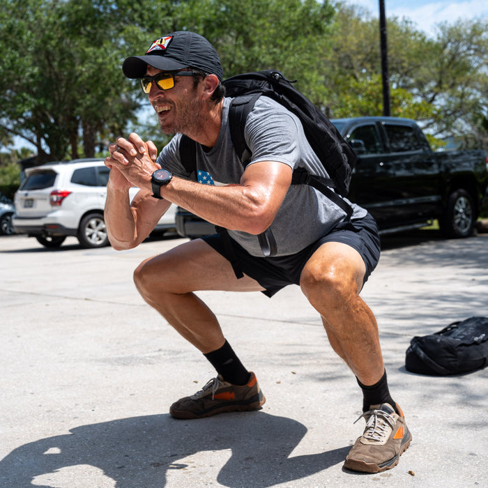 A man in athletic gear does squats outdoors, smiling with a backpack on, while wearing Men’s Training Shorts - ToughStretch for all-day comfort.