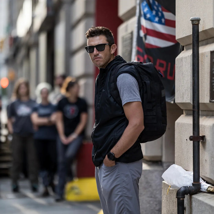 Man wearing GORUCK backpack and sunglasses standing on city street, American flag in background
