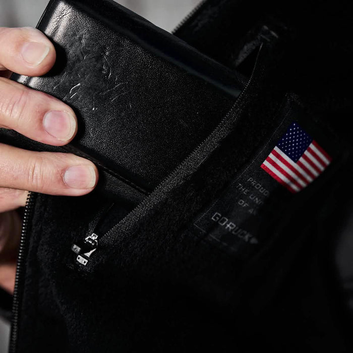 Hand placing black wallet in GORUCK jacket pocket with USA flag patch visible
