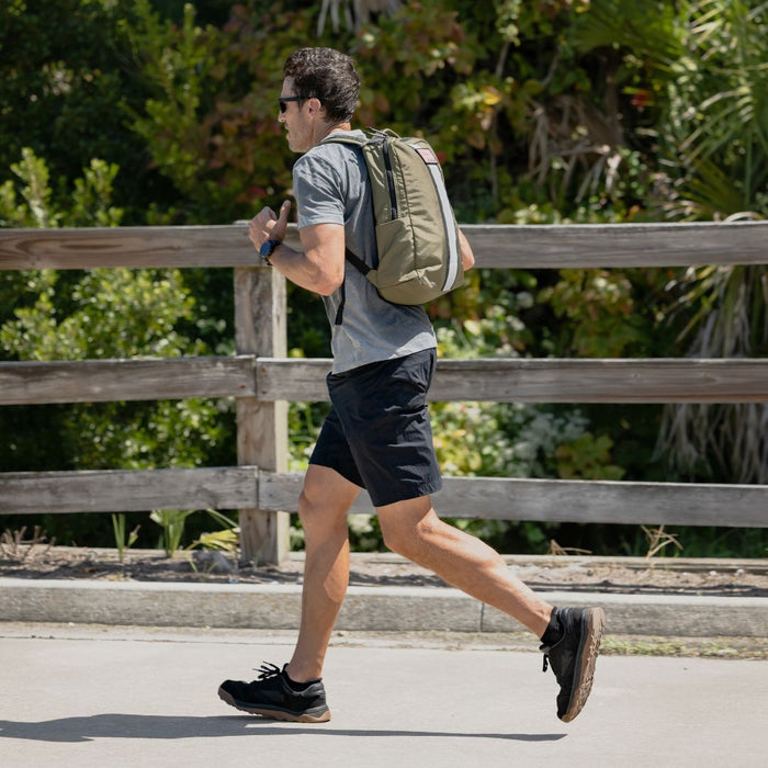 A man jogs on a sunny day, wearing sunglasses, a backpack, athletic clothes, and comfortable Merino Challenge Socks - Ankle (No-Show), with greenery in the background.