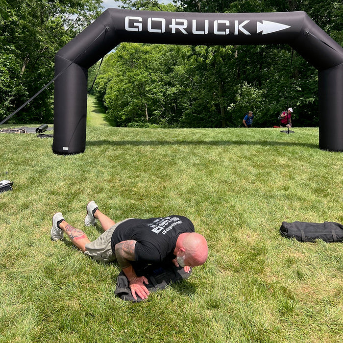 A man wearing Merino Challenge Socks - Ankle (No-Show) does push-ups on grass beneath a “GORUCK” inflatable arch at an outdoor event.