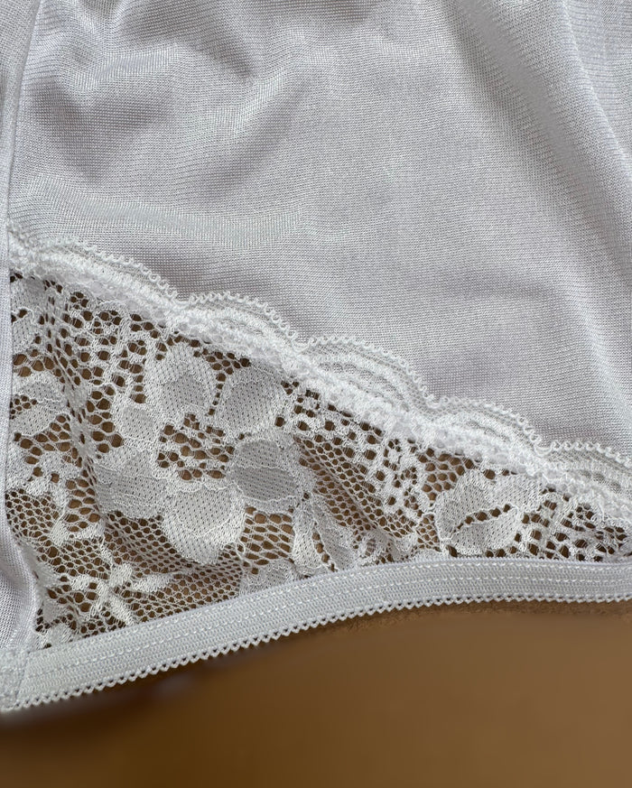 A close-up of white fabric with floral lace trim, like the detailing on Teri Lingerie’s Nylon and Lace Brief Panty (Style M108/Beatrice) 3 Pack in White, displayed on a beige surface.