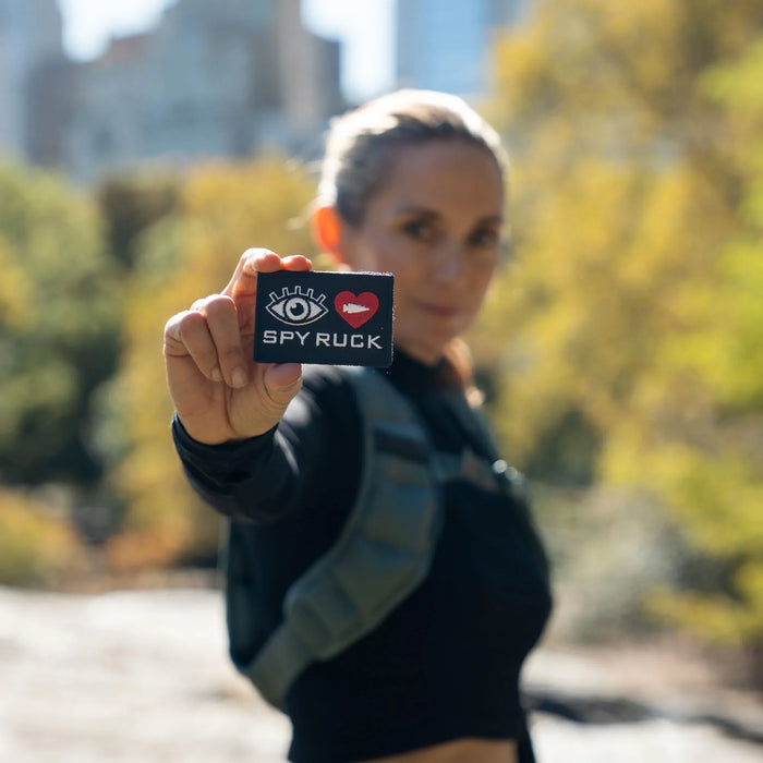 Woman outdoors with GORUCK rucksack holds SPY RUCK patch in focus, blurred background.