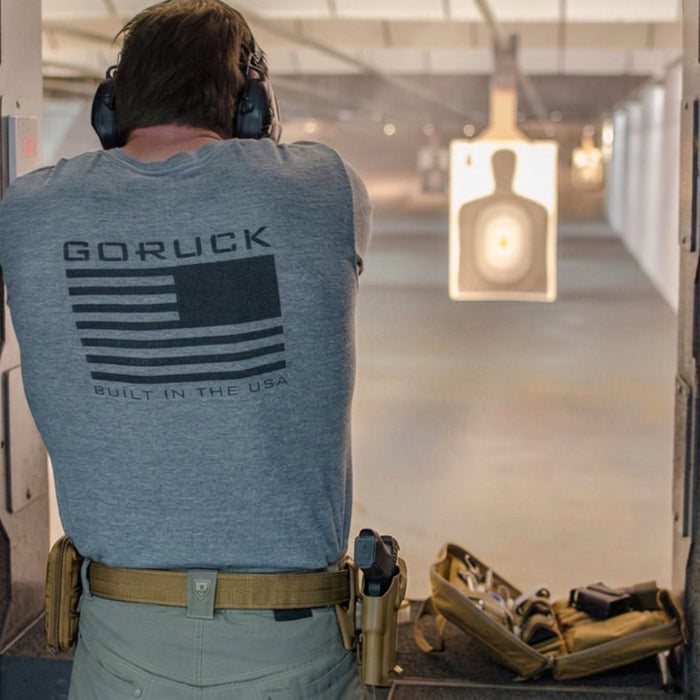 At an indoor shooting range, a person wearing ear protection and a "GORUCK" shirt aims at a paper target, with a Pistol Rug and magazine pouch set on the bench beside them.