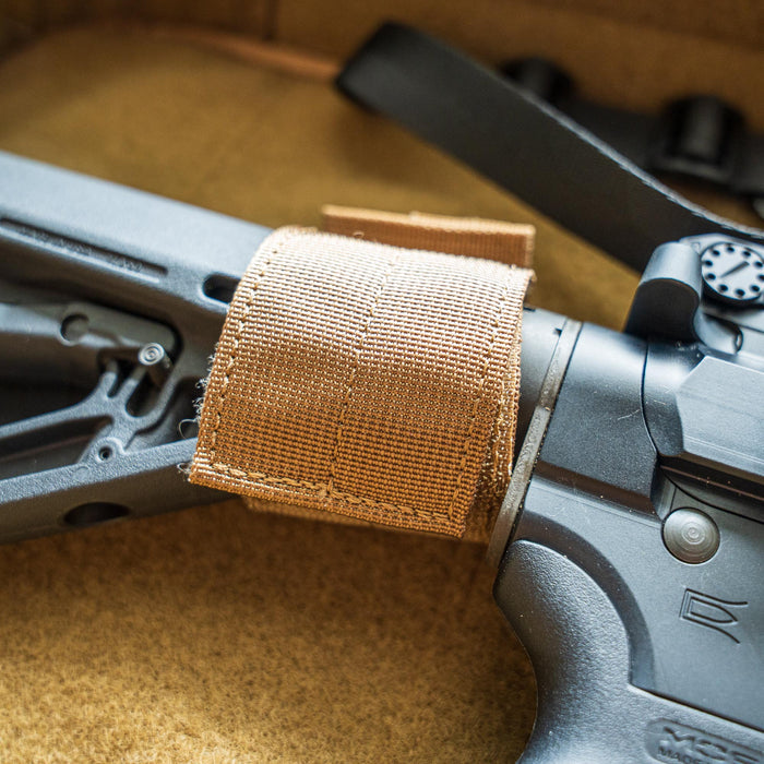 Close-up of the Retention Package's tan nylon strap securing a black firearm stock on a tan surface.