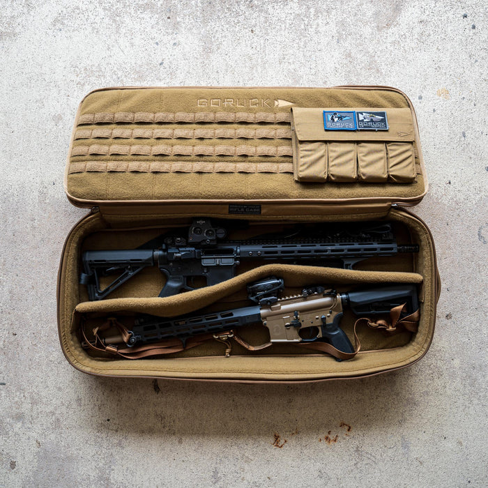 A Rifle Case in tan, adorned with patches, lies open on a concrete floor, displaying two rifles inside.