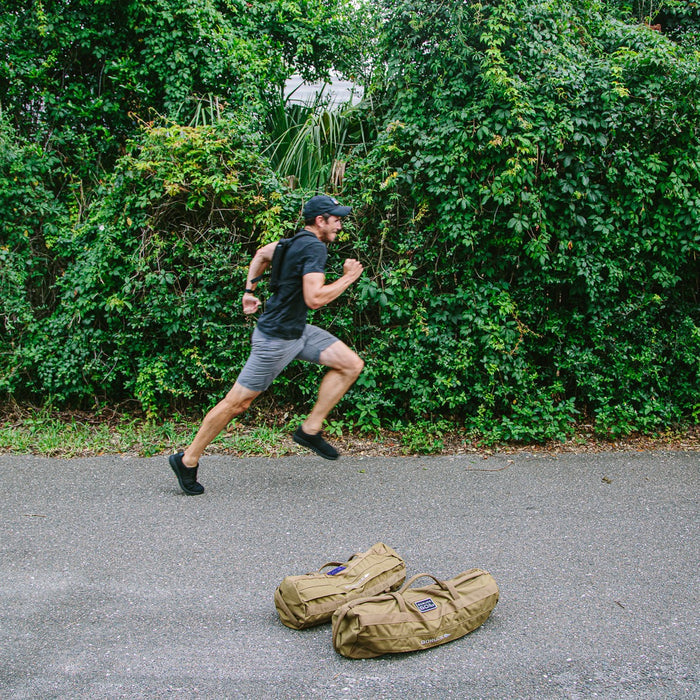 A man in athletic wear runs past two duffel bags on a paved path, wearing the Ruck Plate Carrier 3.0 for enhanced lumbar support, with dense greenery in the background.
