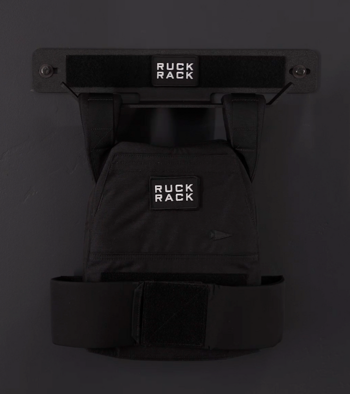 A black Ruck Rack® Delta | Heavy-Duty Wall Mount Storage for Rucks, Weight Vests & Tactical Gear holds a black rucksack labeled “Ruck Rack,” offering a sleek and practical wall-mounted solution for organizing plate carriers and other tactical gear.