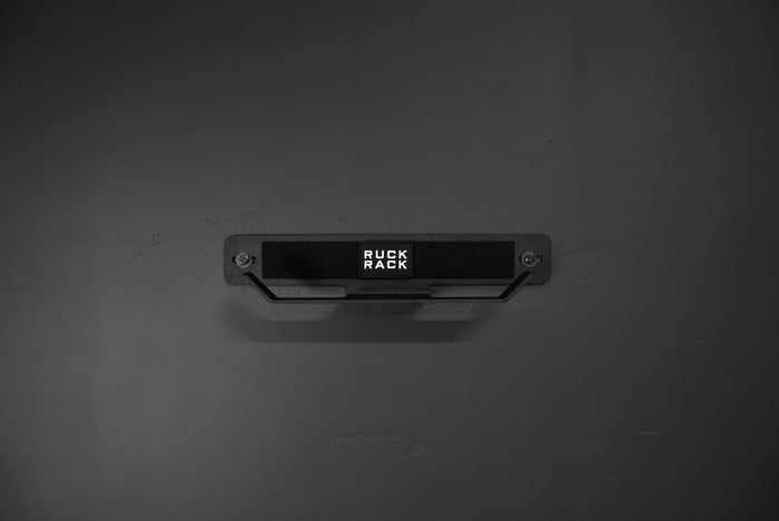 A Ruck Rack® Delta, a heavy-duty black wall-mounted storage rack for rucks, weight vests, and tactical gear, is centered on a dark wall, serving both as a versatile organizer and practical display solution.