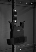 A black Ruck Rack® Monster, a versatile mount for rucks, weight vests, and tactical gear, hangs on a metal gym rack in a dimly lit room—serving as the perfect organizer for your training sessions.