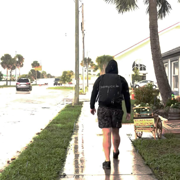 Person rucking in rain with GORUCK black rain cover on backpack, walking on wet sidewalk