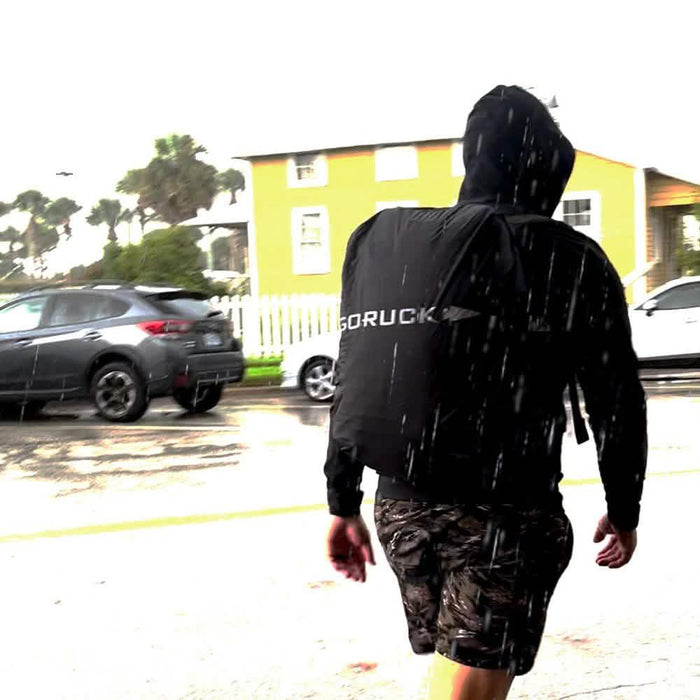 Person wearing GORUCK rain cover on backpack in heavy rain, outdoor urban setting