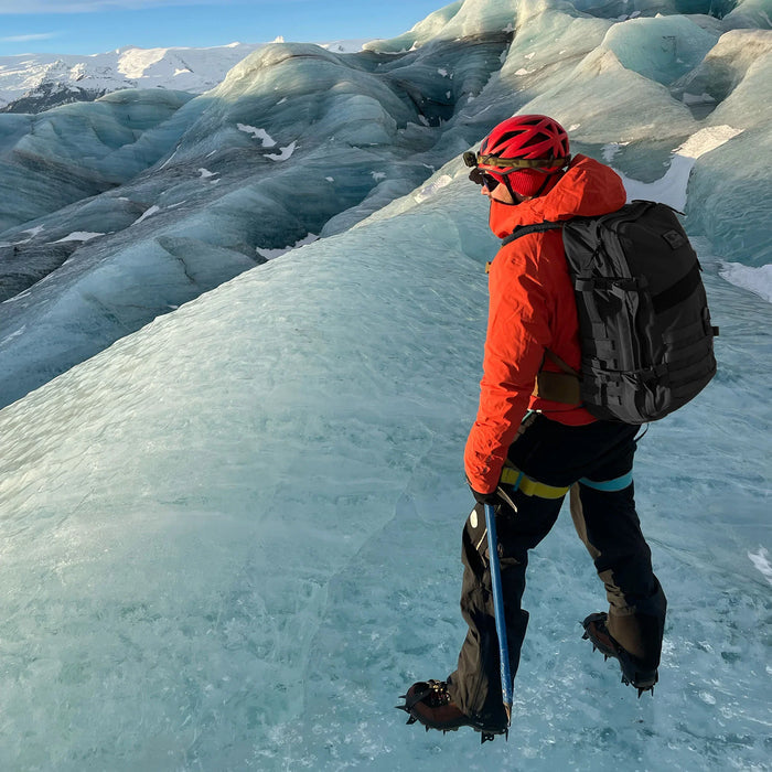 Mountaineer in red jacket with GORUCK backpack climbing icy glacier, wearing crampons and helmet