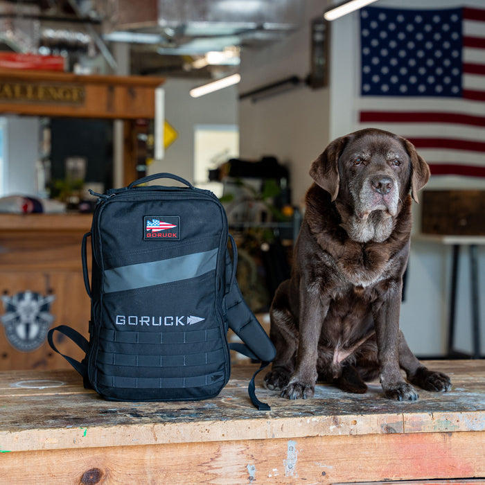 A brown dog sits beside a black Rucker 5.0 rucksack on a wooden table, with a US flag in the background, ready for adventure.