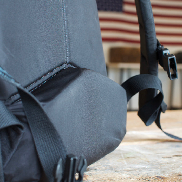 Close-up of the Rucker 5.0, a black rucksack with lumbar support, resting on a wooden surface with an American flag blurred in the background.
