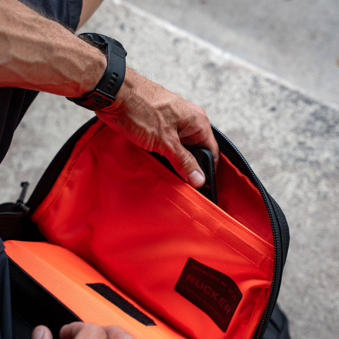 A person with a black watch puts a phone into an orange-lined pocket of the Rucker 5.0 rucksack, designed with lumbar support for comfortable rucking.
