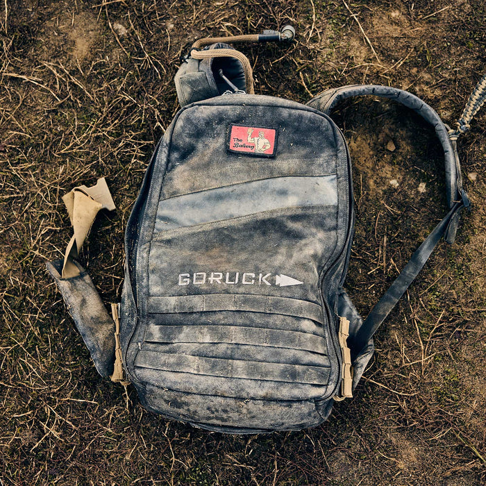 A dusty, worn Rucker 5.0 rucksack with a red patch on top rests on dry, patchy ground—ready for another day of rucking and providing solid lumbar support.